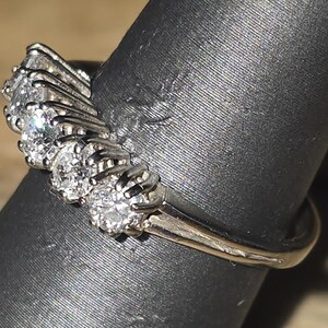May include: A silver ring with multiple clear, round-cut gemstones arranged in a curved design. The ring is set on a black surface, showcasing the sparkle of the stones and the metal's shine.