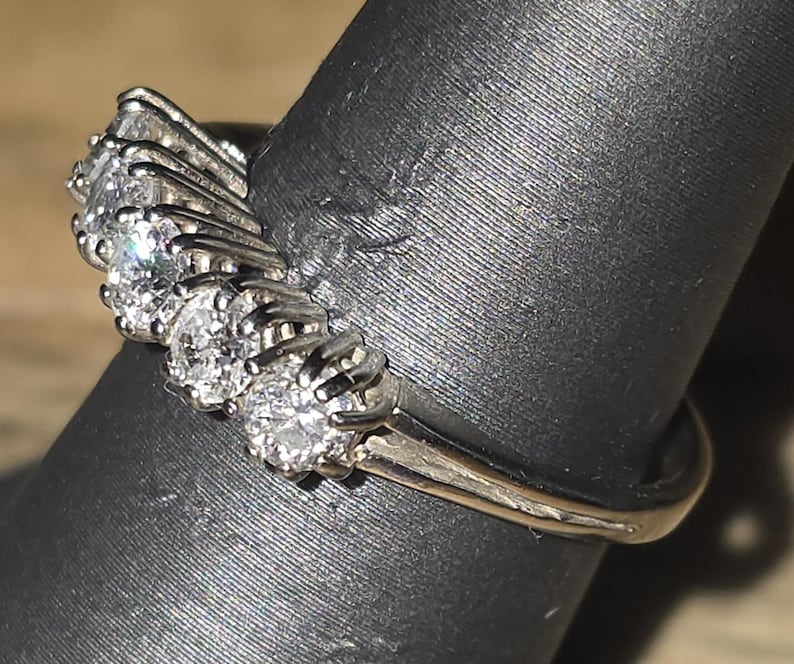 May include: A silver ring with multiple clear, round-cut gemstones arranged in a curved design. The ring is set on a black surface, showcasing the sparkle of the stones and the metal's shine.