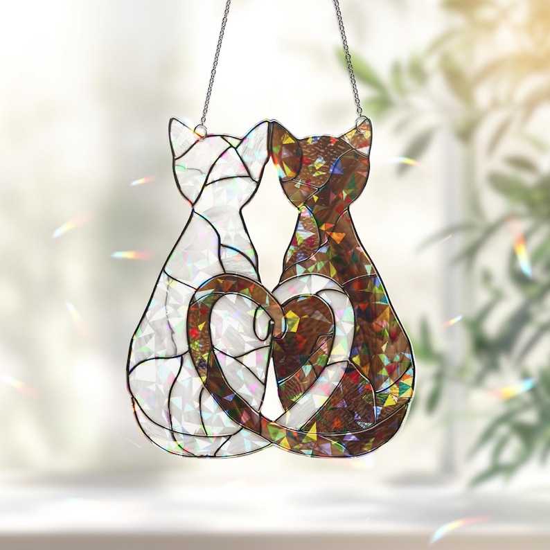 Puede incluir: Atrapasoles de gato de vidriera con dos siluetas de gato formando un coraz&oacute;n. Un gato es blanco, el otro marr&oacute;n con detalles iridiscentes. La pieza cuelga de una cadena, ideal para una ventana o pared.