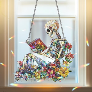 May include: Stained glass window hanging featuring a skeleton reading a book while seated among colorful flowers. The skeleton and flowers are made of various colors of glass, and the piece hangs from a chain.