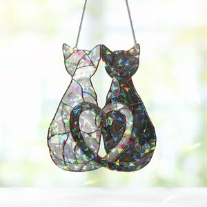 Puede incluir: Un atrapasoles de cristal con dos gatos cuyas colas forman un coraz&oacute;n. Un gato es plateado y el otro negro, ambos con detalles iridiscentes. El atrapasoles cuelga de una cadena.