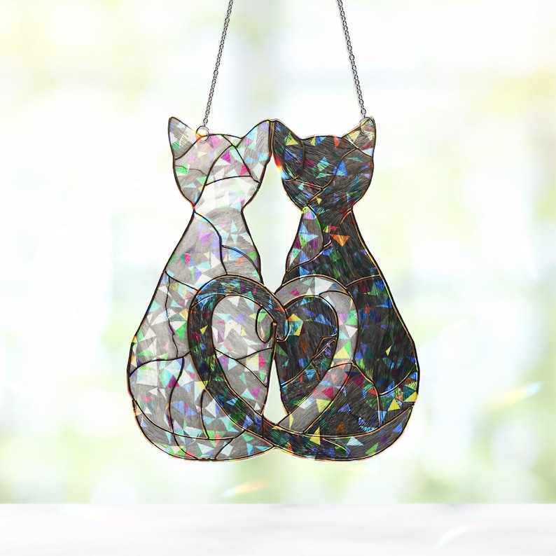 Puede incluir: Un atrapasoles de cristal con dos gatos cuyas colas forman un coraz&oacute;n. Un gato es plateado y el otro negro, ambos con detalles iridiscentes. El atrapasoles cuelga de una cadena.