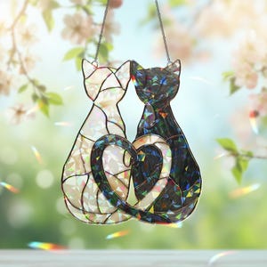 Puede incluir: Un atrapasoles de vidrieras que muestra dos gatos sentados uno al lado del otro. Un gato es blanco y el otro es negro, ambos con una textura facetada e iridiscente. Sus colas se curvan juntas para formar un coraz&oacute;n. La pieza cuelga de una cadena fina.