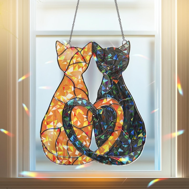 Puede incluir: Decoraci&oacute;n de ventana de vidriera con dos siluetas de gatos enfrentadas, formando un coraz&oacute;n con sus colas. Un gato es naranja, el otro negro, ambos con patrones geom&eacute;tricos. Cuelga de una cadena.