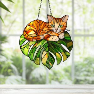 May include: Stained glass window hanging featuring an orange tabby cat resting on a green monstera leaf.