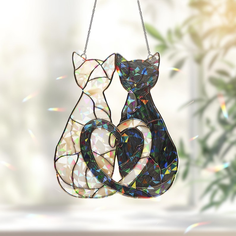 Puede incluir: Atrapasoles de gato de vidriera con dos gatos cuyas colas forman un coraz&oacute;n. Un gato es blanco, el otro negro, ambos con detalles iridiscentes. Los gatos est&aacute;n suspendidos por una cadena plateada.