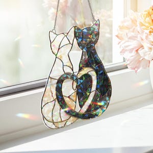 Puede incluir: Atrapasoles de gato de vidriera con dos gatos con colas entrelazadas formando un coraz&oacute;n. Un gato es blanco, el otro negro e iridiscente. El atrapasoles cuelga de una cadena, creando reflejos de arco&iacute;ris.