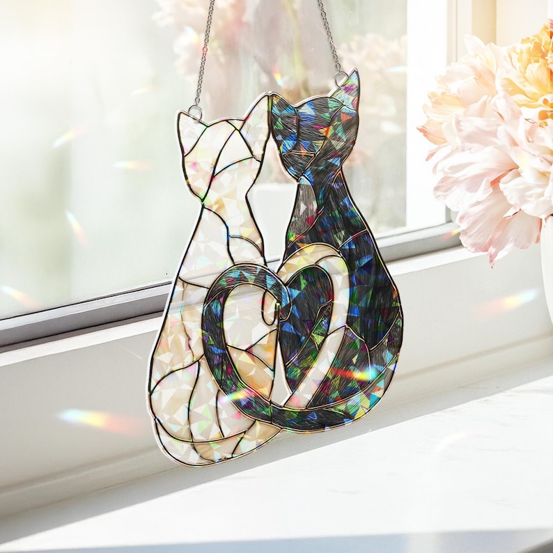 Puede incluir: Atrapasoles de gato de vidriera con dos gatos con colas entrelazadas formando un coraz&oacute;n. Un gato es blanco, el otro negro e iridiscente. El atrapasoles cuelga de una cadena, creando reflejos de arco&iacute;ris.