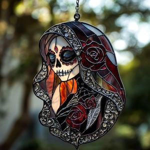 May include: Stained glass art piece depicting a stylized skull with floral and ornamental details. The piece features a red rose, a feather, and a decorative hood. The color palette includes red, black, and white, with a chain for hanging.