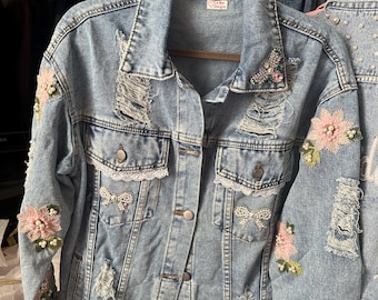 May include: Light blue denim jacket with distressed details, adorned with embroidered pink flowers, bows, and pearl accents. Features a classic collar, button-up front, and chest pockets. The jacket has a label that says "La Mar Designs".