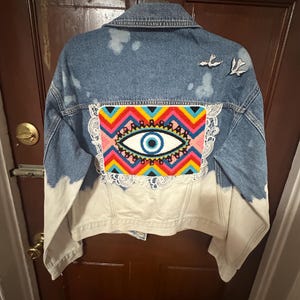 May include: A denim jacket with a colorful embroidered eye design on the back. The jacket features a light blue wash with white accents and a lace trim around the embroidered patch. Two small bird patches are on the upper back.