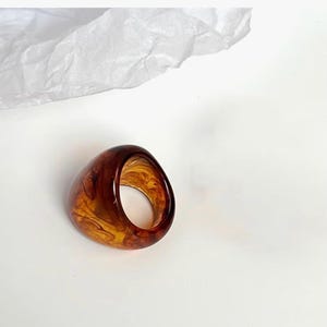 May include: A chunky ring crafted from translucent resin featuring a swirling pattern of deep amber and golden brown tones. The ring has a smooth, rounded, and thick band design with a polished finish that catches the light, creating depth.