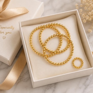 Peut inclure: Trois bracelets en perles dorées et une bague en perles assortie disposés dans une boîte cadeau blanche. Le couvercle de la boîte présente un ruban doré et le texte Especially for you avec un petit symbole de cœur imprimé à l'encre dorée.