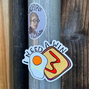 May include: A sticker with a cartoon illustration of a fried egg and a slice of toast with the text "I need a win" in black lettering.