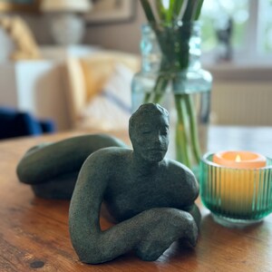 May include: A dark green ceramic sculpture of a person lying down with their arms crossed. The sculpture is on a wooden table with a green glass candle holder and a lit candle.