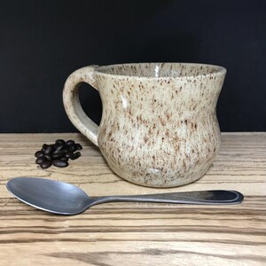 May include: A speckled beige ceramic mug with a brown handle. The mug is sitting on a wooden surface with a spoon and coffee beans.