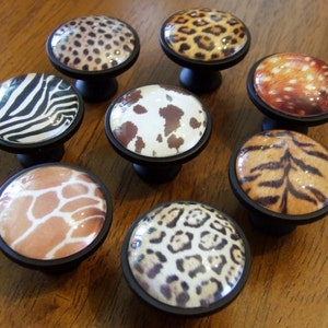 May include: A collection of eight decorative cabinet knobs with animal print designs. The knobs feature patterns like leopard, zebra, cowhide, giraffe, and tiger. Each knob has a black base and a glossy, rounded top, offering a unique touch for furniture.