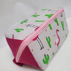 May include: A rectangular cosmetic bag with a pink zip and accents. The bag features a light grey background with a pattern of green cacti, pink flamingos, and gold dots. The bottom and sides are a solid magenta colour.