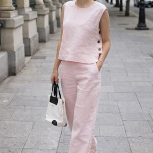 以下が含まれることがあります： ボタン付きノースリーブトップスとワイドパンツのライトピンクのリネンセットアップ。白いフラットシューズと黒のアクセントのトートバッグが組み合わされています。暖かい季節に最適です。