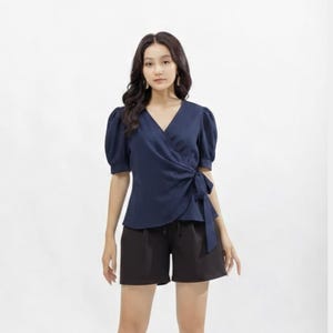 May include: A navy blue wrap top with short, puffed sleeves and a matching tie closure. Paired with black shorts and black high-heeled sandals. The outfit is set against a plain white background.