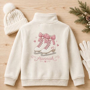 May include: A cream-colored fleece jacket featuring an embroidered design of two white ice skates with pink laces and bows, surrounded by small pink hearts. The name Hannah is embroidered in pink script below the skates, accompanied by a matching beanie.