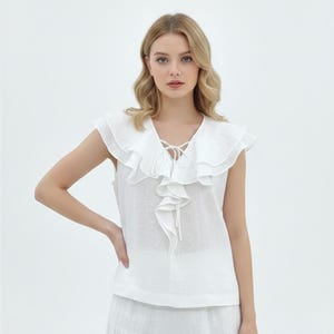 Op de afbeelding: Een witte mouwloze top met een gerimpelde kraag en een V-hals. De top heeft een strikdetail bij de halslijn en wordt gecombineerd met een witte rok. De outfit is gemaakt van een lichte stof, perfect voor warm weer.
