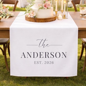 May include: A three-tiered white wedding cake with pink and white flowers on a wooden base. The cake is on a table with a white runner that reads "The ANDERSON EST. 2026". The table is set for a wedding reception.