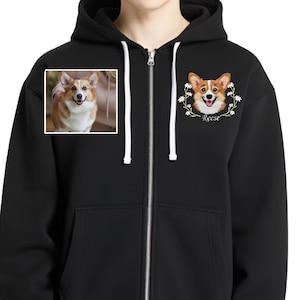 May include: Black zip-up hoodie featuring two corgi dog portraits. One is a photo in a square frame, the other is an illustration with the name "Reese" below. The hoodie has a hood and a full-length zipper.