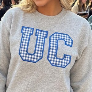Könnte beinhalten: Ein graues Sweatshirt mit den Buchstaben "UC" in blau-weißem Vichy-Muster. Das Sweatshirt wird von einer Person mit blonden Haaren getragen, die vor einem Backsteingebäude und einer Gruppe von Menschen lächelt.