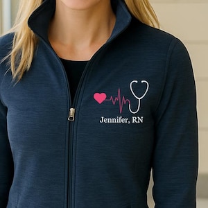 Puede incluir: Chaqueta azul marino con cremallera y un gráfico de estetoscopio y latido cardíaco en rosa y blanco. El texto "Jennifer, RN" está impreso debajo. La chaqueta tiene cuello alto y dos bolsillos delanteros.