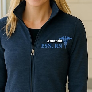 May include: Navy blue zip-up jacket with the name "Amanda", the initials "BSN, RN", and a medical caduceus symbol embroidered in blue. The jacket has a high collar and two front pockets.