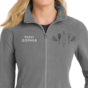 May include: A gray fleece zip-up jacket with a high collar. The jacket features the text "Baker SOPHIA" and a graphic of baking tools. The jacket has two front pockets and a silver zipper.