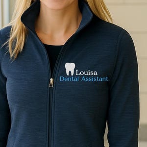 May include: Navy blue zip-up jacket with the text "Louisa Dental Assistant" and a tooth graphic. The jacket has a stand-up collar, long sleeves, and two front pockets. The model is wearing blue jeans.