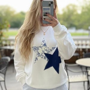 May include: A white sweatshirt featuring a large star design. The star is a combination of a dark blue star shape with a floral pattern in the centre. The person is also wearing blue and white striped shorts.