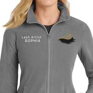May include: A gray fleece jacket with a full zipper and two pockets. The jacket has the text "Lash Artist SOPHIA" on the left chest and a black eyelash design with gold stars on the right chest.