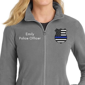 Puede incluir: Chaqueta de forro polar gris con cremallera completa, dos bolsillos y cuello alto. La chaqueta tiene el texto "Emily Police Officer" en el pecho izquierdo y una insignia de policía con una línea azul en el derecho.
