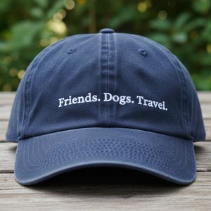 May include: Navy blue baseball cap with the embroidered text "Friends. Dogs. Travel." The hat is made of a soft material and has a curved brim. The hat is sitting on a wooden surface.