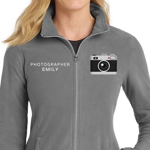 May include: Grey fleece jacket with a full zip, two pockets, and a high collar. The jacket has the text "PHOTOGRAPHER EMILY" and a camera graphic on the chest.