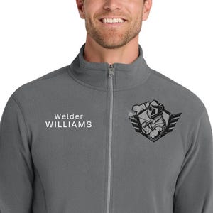 May include: A gray zip-up jacket with the text "Welder WILLIAMS" on the left chest and a black and gray graphic of a welder on the right chest. The jacket has a full zipper and two side pockets.