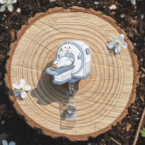 Op de afbeelding: Een wit en blauw badge reel met een cartoon eend in een MRI-machine. De badge reel ligt op een houten schijf omringd door kleine witte bloemen en groen. De reel heeft een doorzichtig intrekbaar plastic koord en een metalen clip.