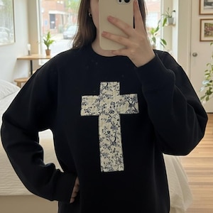 Könnte beinhalten: Schwarzes Sweatshirt mit einem großen Kreuzdesign auf der Vorderseite. Das Kreuz besteht aus weißem Stoff mit einem blauen Blumenmuster. Das Sweatshirt hat lange Ärmel und einen Rundhalsausschnitt.