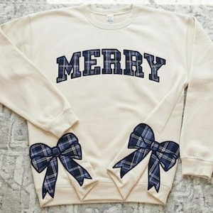 Pode incluir: Camisola creme com a palavra "MERRY" em letras xadrez azul marinho no peito. Apresenta dois laços xadrez azul marinho, um em cada manga. Uma peça festiva e aconchegante com tema de feriado.