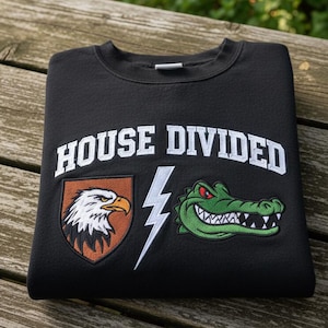 Könnte beinhalten: Schwarzes Sweatshirt mit dem weißen Schriftzug "HOUSE DIVIDED". Das Design umfasst ein Adlerwappen, einen Blitz und eine grüne Alligatorengrafik. Das Sweatshirt liegt gefaltet auf einer Holzoberfläche.