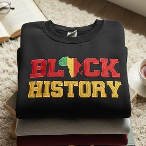 May include: A black sweatshirt featuring the embroidered text "BLACK HISTORY" in red and gold, with a map of Africa in green, yellow, and red. The sweatshirt is stacked with other folded sweatshirts. A cup of tea and an open book are in the background.