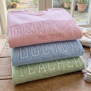Może przedstawiać: Trzy bluzy w pastelowych kolorach ułożone na drewnianej powierzchni. Górna bluza jest różowa i ma napis "DENTIST Est. 2026". Poniżej znajduje się niebieska bluza z napisem "DOCTOR Est. 2026". Dolna bluza jest zielona i ma napis "TEACHER Est. 2026".