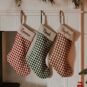 Puede incluir: Tres calcetines navideños colgantes con un patrón de cuadros vichy en rojo y verde. Cada calcetín tiene un puño beige bordado con los nombres Emma, Noah y Sophie en letra cursiva. Los calcetines cuelgan de una repisa de chimenea blanca.
