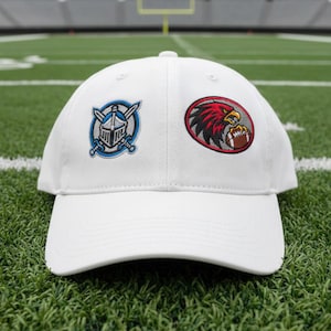 May include: White baseball cap with two embroidered patches. One patch features a blue knight's helmet and crossed swords, the other a red eagle clutching a football. The cap is on a green field.