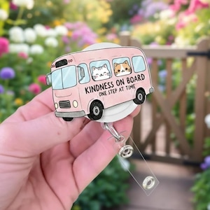 Può includere: Un porta badge a forma di autobus rosa con la scritta "KINDNESS ON BOARD ONE STEP AT TIME". L'autobus ha due gatti passeggeri. Il porta badge ha un cinturino in plastica trasparente e una clip in metallo.
