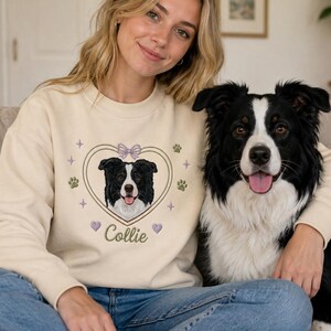 May include: A beige crewneck sweatshirt featuring an embroidered design of a black and white Border Collie dog centered within a heart outline. The design includes a small purple bow, paw prints, stars, and the word Collie in cursive script.