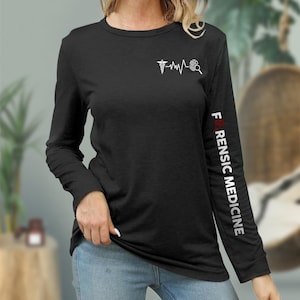 Könnte beinhalten: Schwarzes Langarmshirt mit einer weißen Grafik eines medizinischen Symbols, eines Herzschlags und einer Lupe. Der rechte Ärmel trägt den Text "I ♥ FORENSIC MEDICINE" in Weiß mit einem roten Herzen.